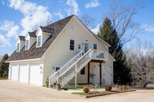A white two-story house with a steep roof, stairs leading to the entrance, and a gravel driveway under a blue sky.