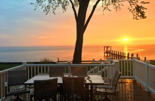 A serene sunset over a calm lake, viewed from a deck with a table and chairs, framed by a large tree.