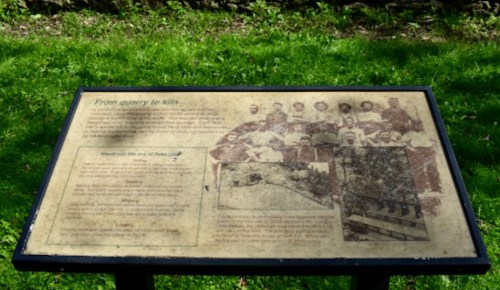 A weathered informational plaque with text and historical photos, set against a green grassy background.