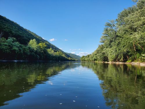 A serene river flows between lush green hills under a clear blue sky, reflecting the landscape in calm waters.