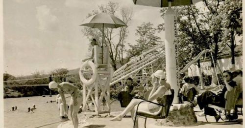 Vintage photo of a swimming pool scene with people relaxing, a lifeguard on duty, and a water slide in the background.