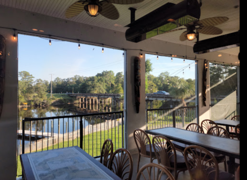 View from a restaurant with tables overlooking a river and a bridge, surrounded by greenery and string lights.