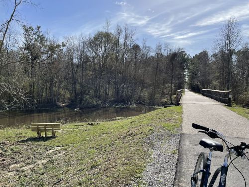 A peaceful scene of a bike path beside a calm river, with trees and a bench in the background under a clear blue sky.