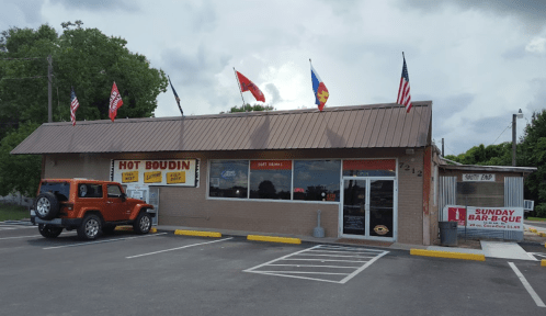 A brick building with a metal roof, featuring flags and a sign for "Hot Boudin," parked Jeep in front.