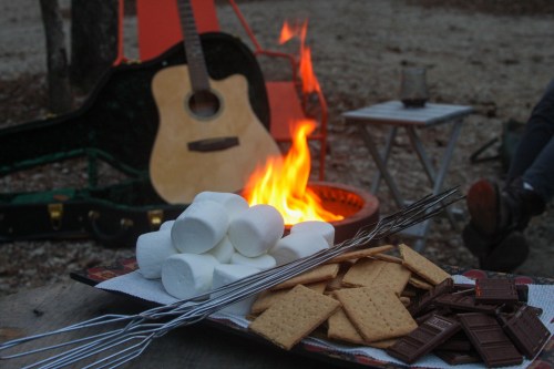 A campfire scene with marshmallows, graham crackers, and chocolate for s'mores, alongside a guitar and camping gear.