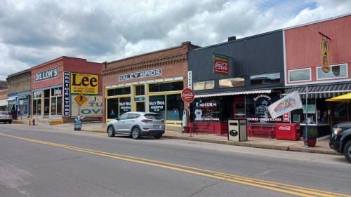 A street view of colorful storefronts, including Lee's and Haley Bros, with a parked car and cloudy sky above.