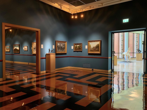 Art gallery interior featuring framed paintings, a sculpture, and a reflective floor with geometric patterns.