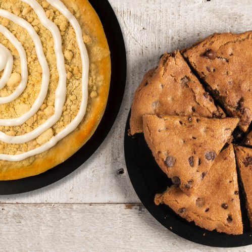 A pie with cream topping and a cookie cake with chocolate chips, both on black plates against a wooden background.