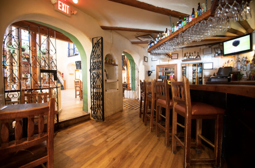 Interior of a cozy restaurant with a bar, wooden furniture, and a warm atmosphere, featuring arches and a well-stocked shelf.