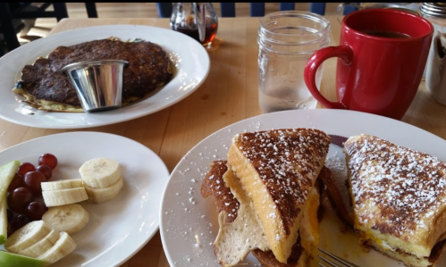 A table with a plate of French toast dusted with powdered sugar, a side of fruit, and a pancake with syrup.