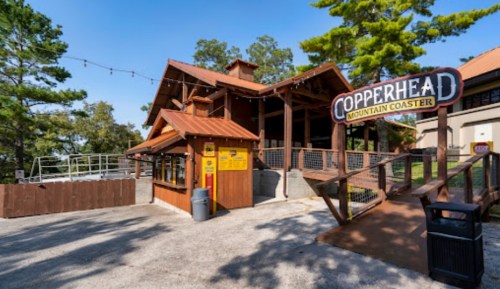 A wooden building with a sign reading "Copperhead Mountain Coaster," surrounded by trees and a gravel path.
