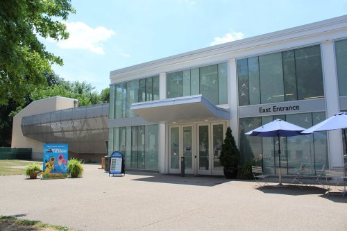 Modern building with large glass windows, featuring an "East Entrance" sign and outdoor seating under blue umbrellas.