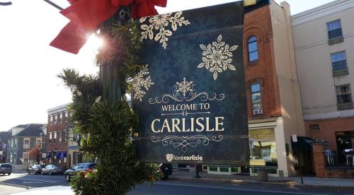 A decorative sign reading "Welcome to Carlisle," adorned with snowflakes and greenery, hangs in a festive setting.
