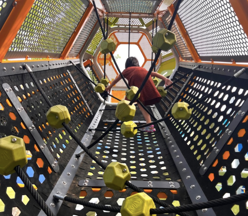 A child climbs through a colorful playground structure with hexagonal patterns and climbing ropes.