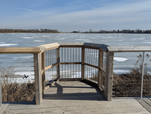 A wooden observation deck overlooking a frozen lake with patches of ice and a clear blue sky.