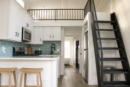 A modern tiny home interior featuring a kitchen, stairs, and a loft area with a railing. Light and airy design.