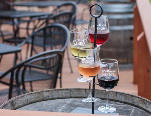 Four glasses of wine in a stand, featuring different colors, on a wooden table at a winery.