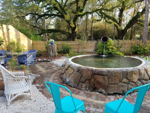 A serene garden with a stone pond, surrounded by greenery, chairs, and a wooden fence in a peaceful setting.