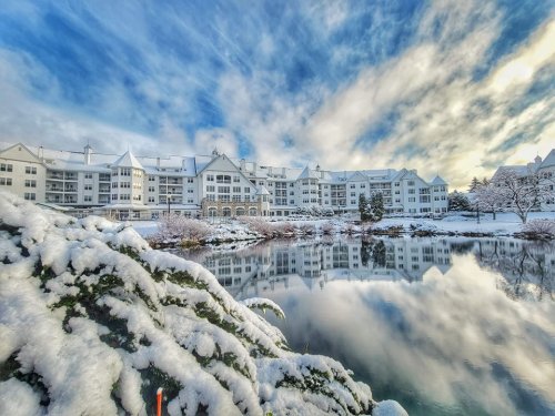 A snowy landscape featuring a lake reflecting a white building and blue sky, surrounded by snow-covered trees.