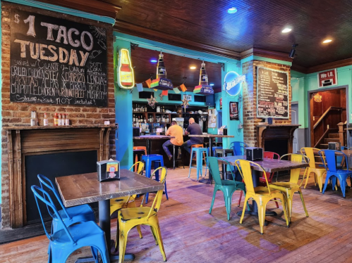 Colorful restaurant interior featuring tables, chairs, and a chalkboard advertising $1 Taco Tuesday. Two patrons at the bar.