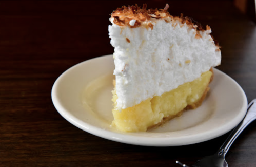 A slice of coconut cream pie on a white plate, featuring a golden filling and a fluffy meringue topping.