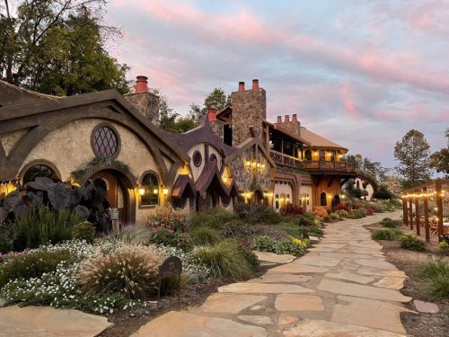 A whimsical, fairy-tale style house with stone and stucco, surrounded by colorful gardens and a sunset sky.