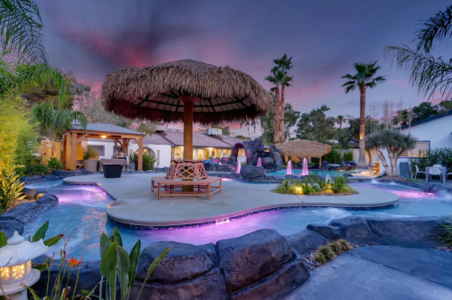 A tropical pool area with a thatched umbrella, palm trees, and colorful lights at sunset.