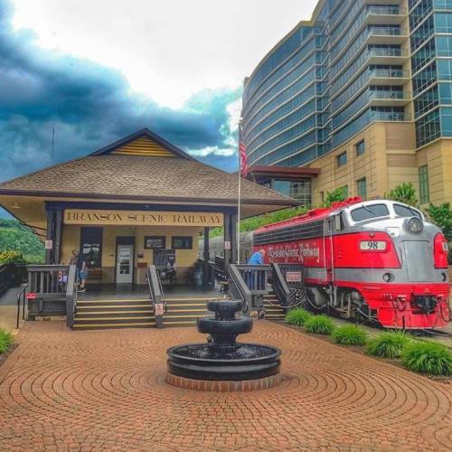 A red train at Branson Scenic Railway station, with a fountain in front and a modern building in the background.