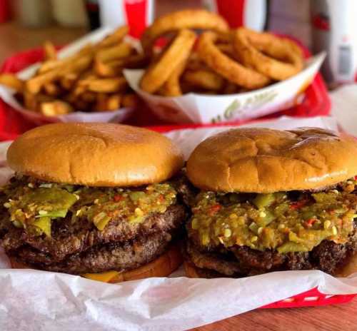 Two juicy burgers with green chili topping, served with a side of fries and onion rings in the background.