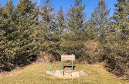 A sign for a nature preserve surrounded by trees and clear blue sky. Green grass in the foreground.