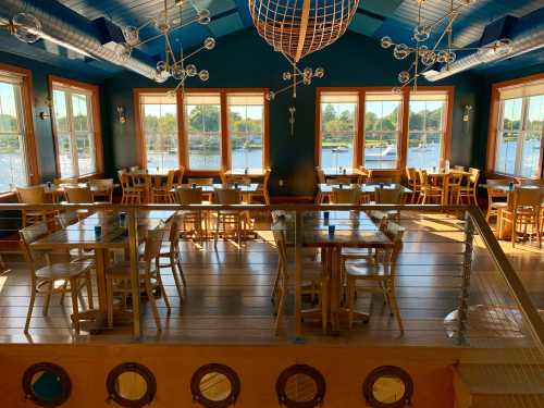 Brightly lit restaurant interior with wooden tables and chairs, large windows overlooking a waterfront view.