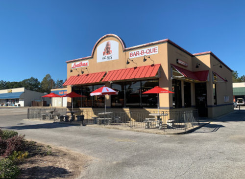 A barbecue restaurant with red umbrellas on the patio, located in a shopping area under a clear blue sky.