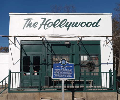 A white building with green trim, featuring "The Hollywood" sign and a historical marker in front.