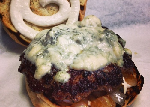 A close-up of a burger with melted blue cheese on a toasted bun, topped with onions and a swirl of sauce.
