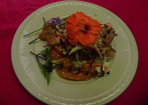 A colorful dish featuring sliced vegetables topped with a vibrant orange flower and garnished with herbs on a plate.