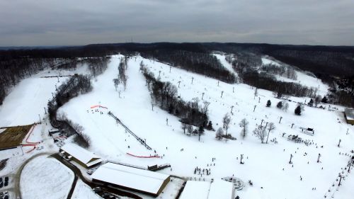 Aerial view of a snowy ski resort with slopes, ski lifts, and scattered skiers under a cloudy sky.