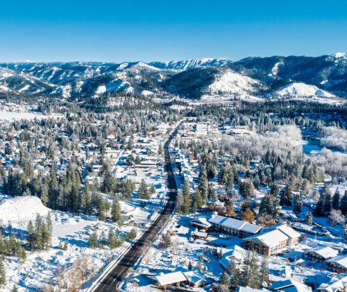 Aerial view of a snowy town surrounded by mountains, with a winding road and trees blanketed in white.