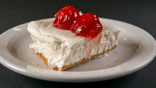 A slice of creamy dessert topped with two cherries on a graham cracker crust, served on a white plate.