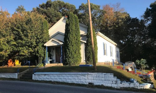 A white house with green trees and shrubs, set on a grassy area beside a road, with colorful play equipment nearby.