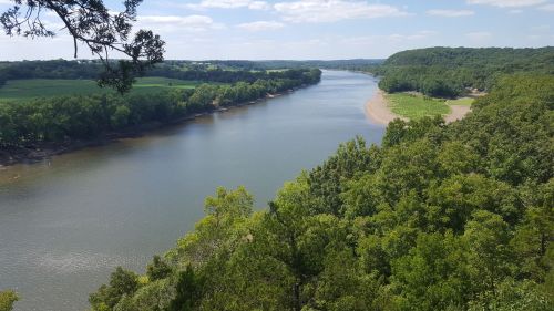 A serene river flows through lush green landscapes under a clear blue sky, surrounded by trees and rolling hills.