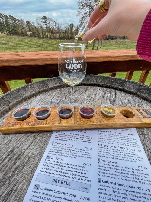 A hand pours wine into a glass beside a tasting flight and menu at Landry Vineyard, with a scenic view in the background.