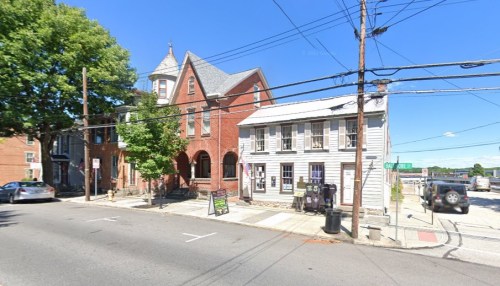 A street view of a red brick building and a white wooden structure, with power lines and parked cars nearby.