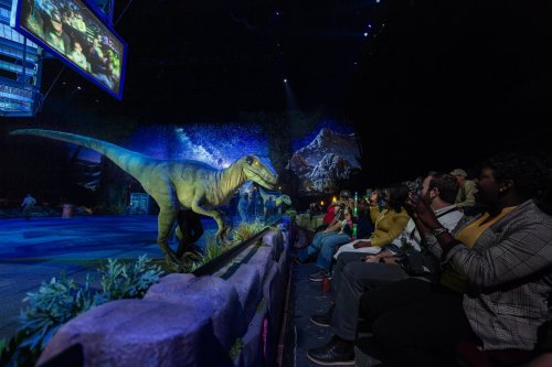 A lifelike dinosaur animatronic interacts with an audience at a live show, with colorful stage lighting in the background.