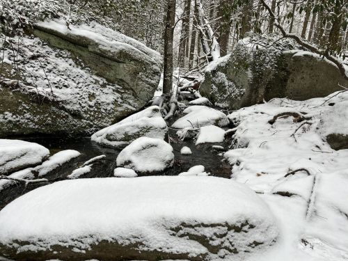 A snowy landscape featuring a stream surrounded by large rocks and trees in a forest setting.