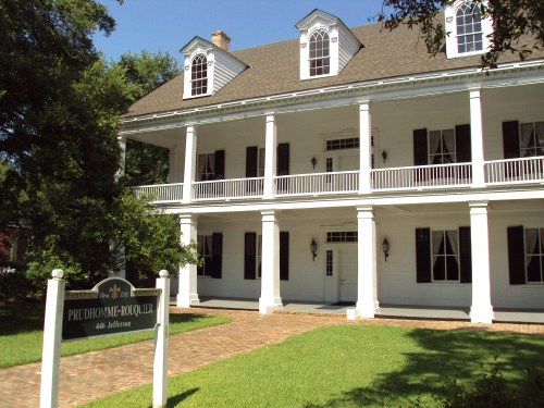 Historic white two-story house with a large porch, surrounded by greenery and a sign reading "PruDhomme-Roquefort."