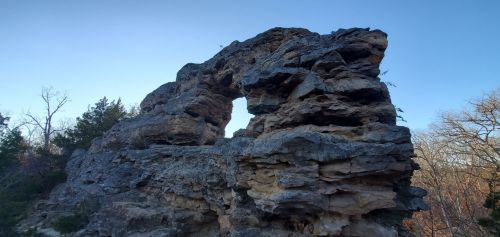A large rock formation with a natural arch, surrounded by trees and a clear blue sky.
