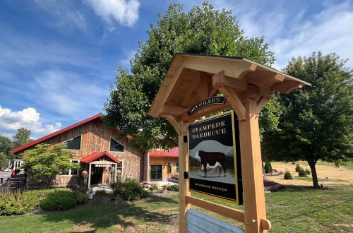 Sign for Stampede Barbecue in front of a rustic building with a red roof, surrounded by trees and blue sky.