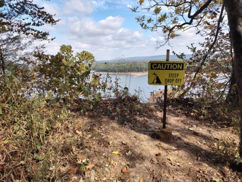 A caution sign warns of a steep drop-off near a river, surrounded by trees and autumn foliage.