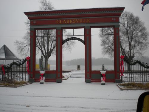 Snow-covered entrance archway to Clarksville, decorated with red bows and flanked by trees in a winter landscape.