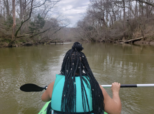 A person with braided hair paddles a kayak on a calm river surrounded by bare trees.